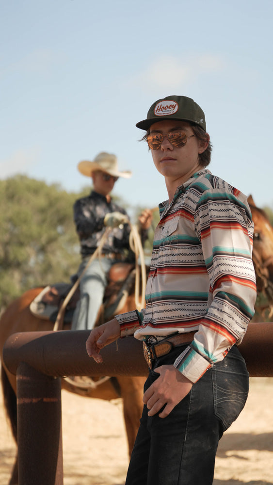 male model posing in dark wash jeans, blue/red/white serape sol shirt and Hooey hat in outdoor setting