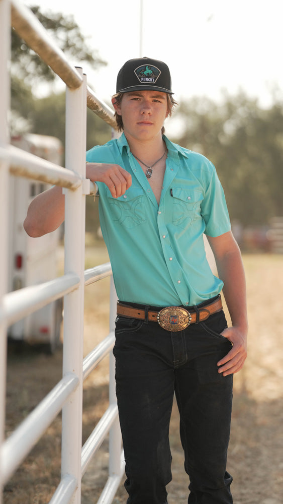 male model wearing light blue sol shirt with dark wash jeans and matching hooey hat, leaning against pipe fence