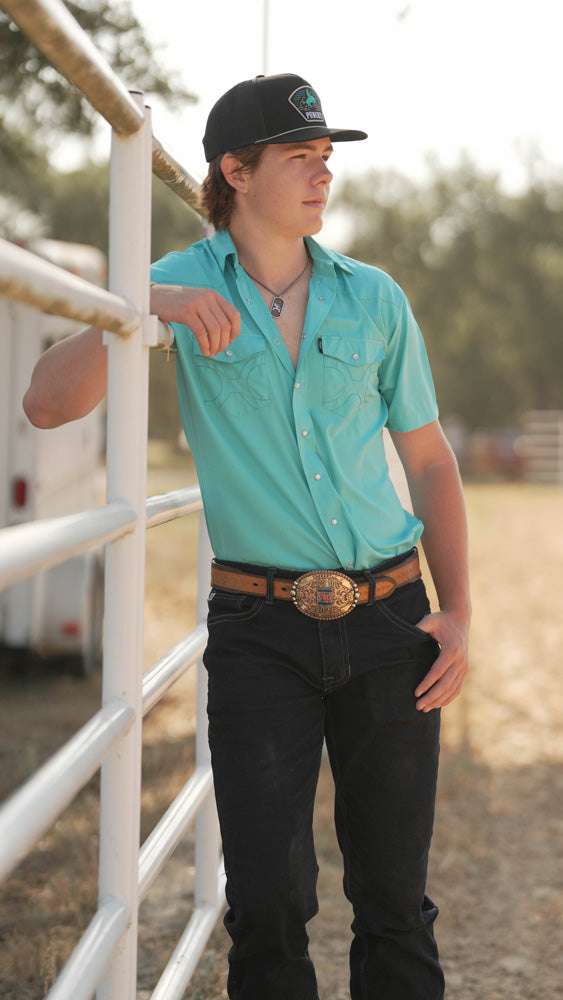 male model wearing black and blue Hooey hat, blue sol shirt, and dark wash jeans while leaning against a pipe fence