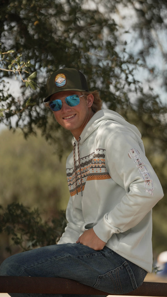 young male model wearing medium wash jeans, white/orange/grey pattern hoody with grey hat and sunglasses posing in outdoor setting