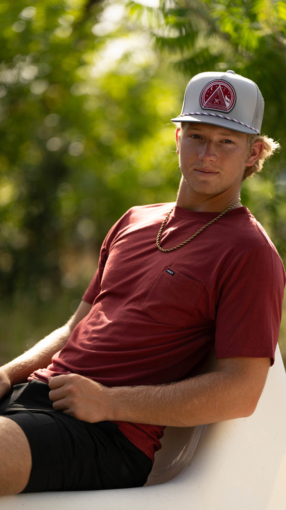 Male model wearing Hooey tee short and board shorts with hooey hat, seated in outdoor setting.