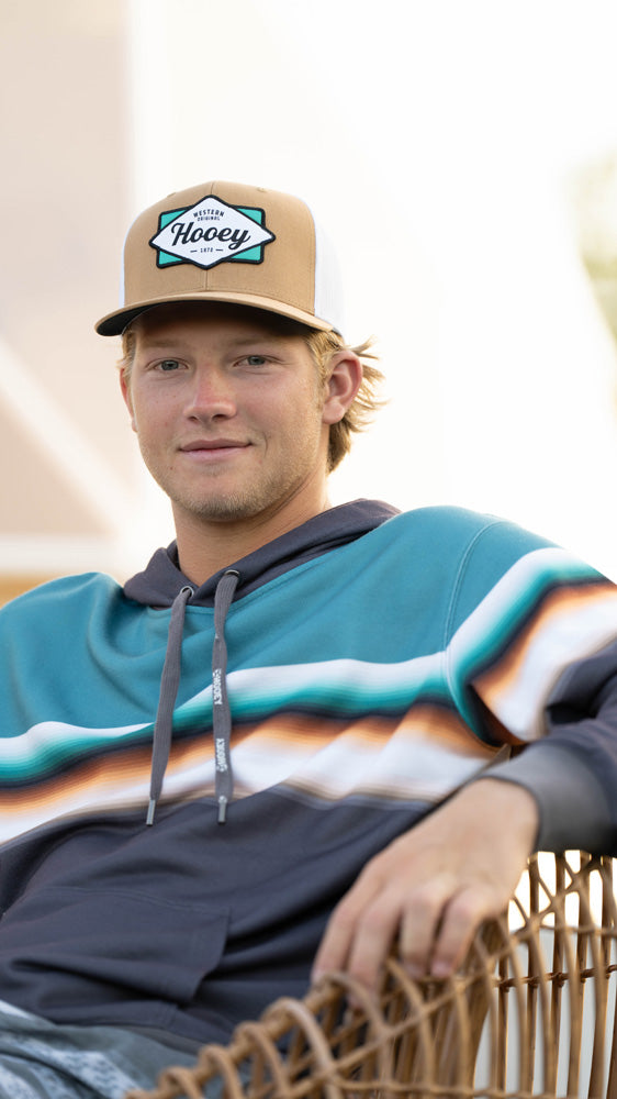 male model wearing tan and white hooey hat with navy/white/orange/light blue stripe pattern hoody in outdoor setting