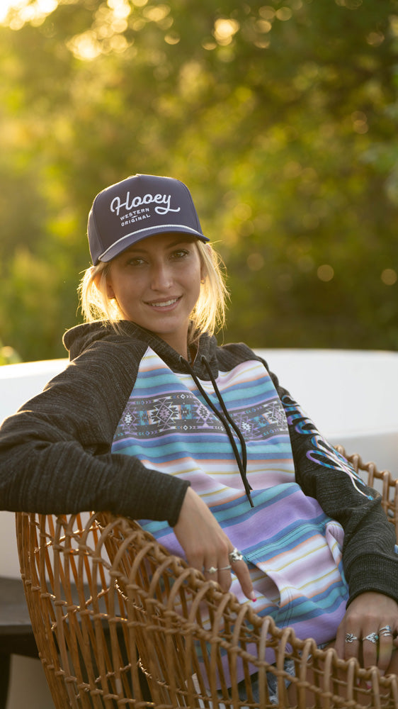 female model wearing maroon Hooey hat with purple and turquoise serape hoody seated in wicker chare in outdoor setting