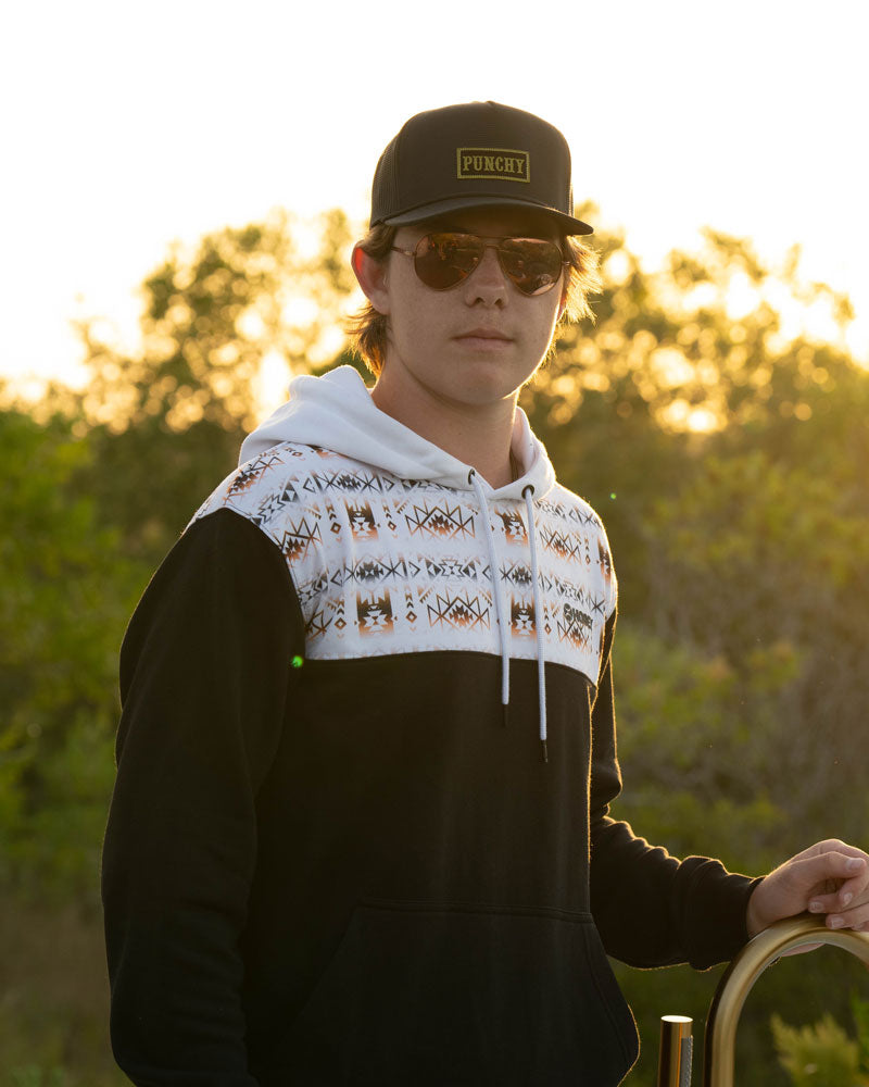 male model wearing black and white Aztec hoody, black and bold at and sunglasses posed in outdoor setting