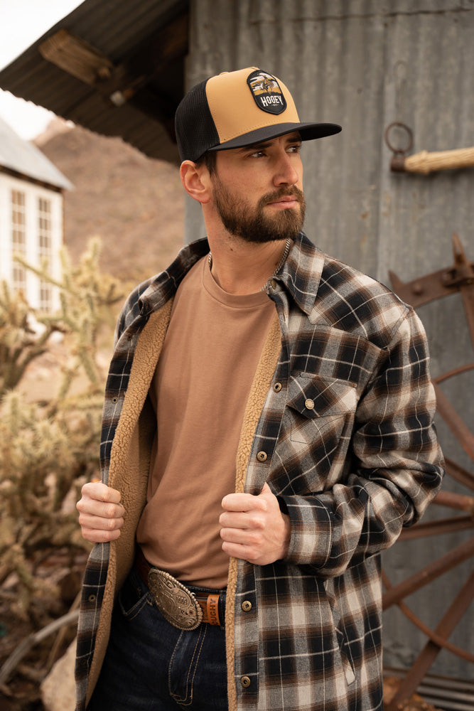 Male model wearing brown T-shirt dark wash jeans brown leather hooey belt brown Gray plaid Shacket with brown fleece lining tan and like hooey Cheyenne had