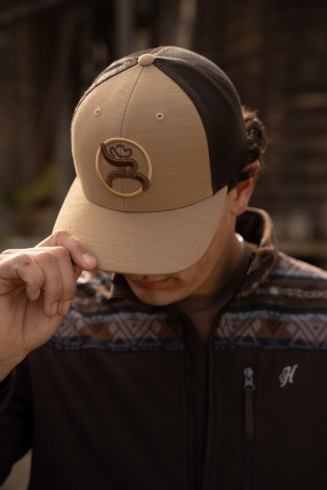 Male model posing and tan and brown fluffy hat with brown tech will zip jacket featuring navy and brown Aztec pattern on collar and shoulders