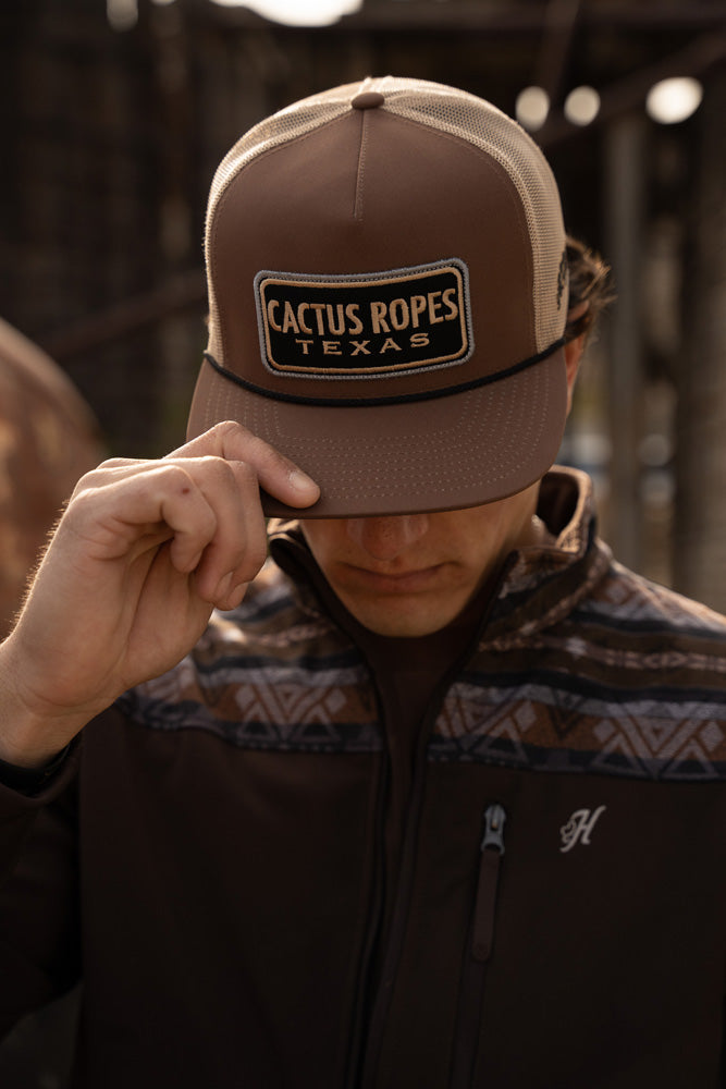 Male model posing with brown and tan cactus Road hand and right hand pulled over eyes looking down Brown and navy Aztec pattern tech jacket