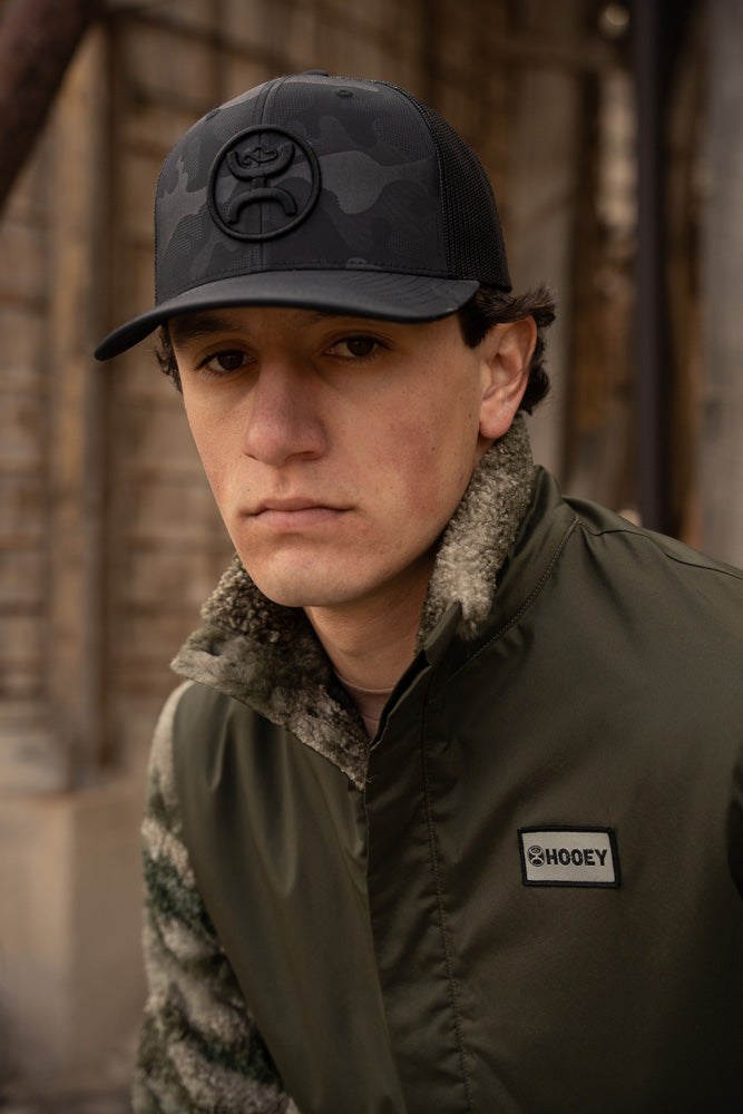 Male model posing in green tech and fleece camo jacket with black and gray camo Hooey hat