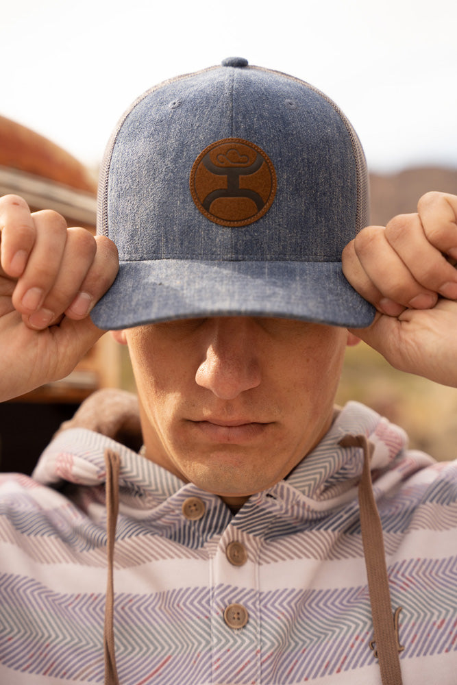 male model wearing tan, white, grey serape hoody with denim and tan hat pulled over eyes with both hands