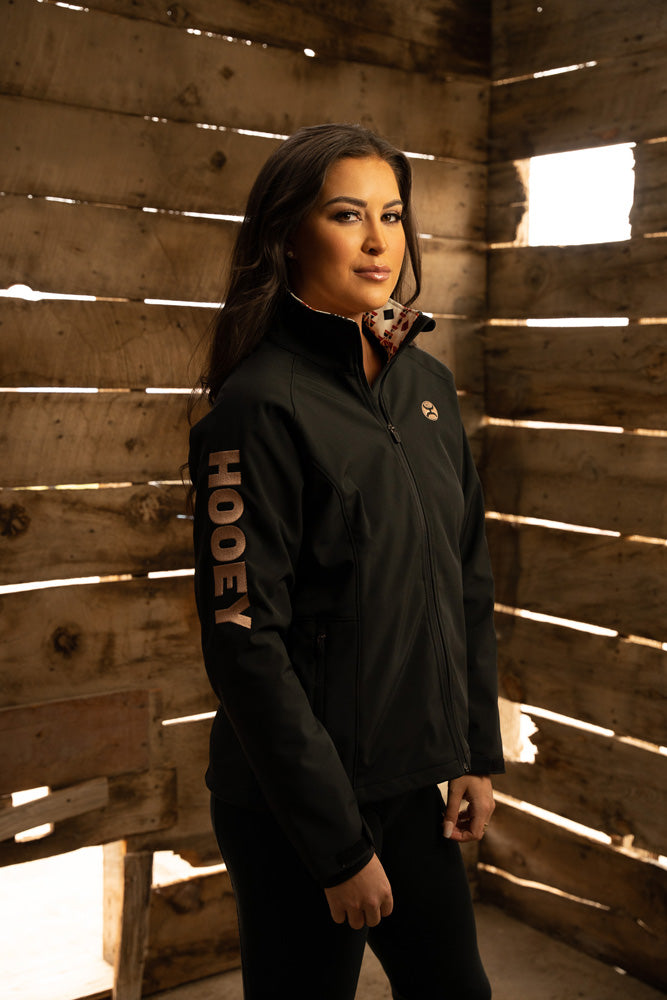 female model wearing black pants and black jacket with pink Hooey logo on the sleeve posed inside a wooden structure