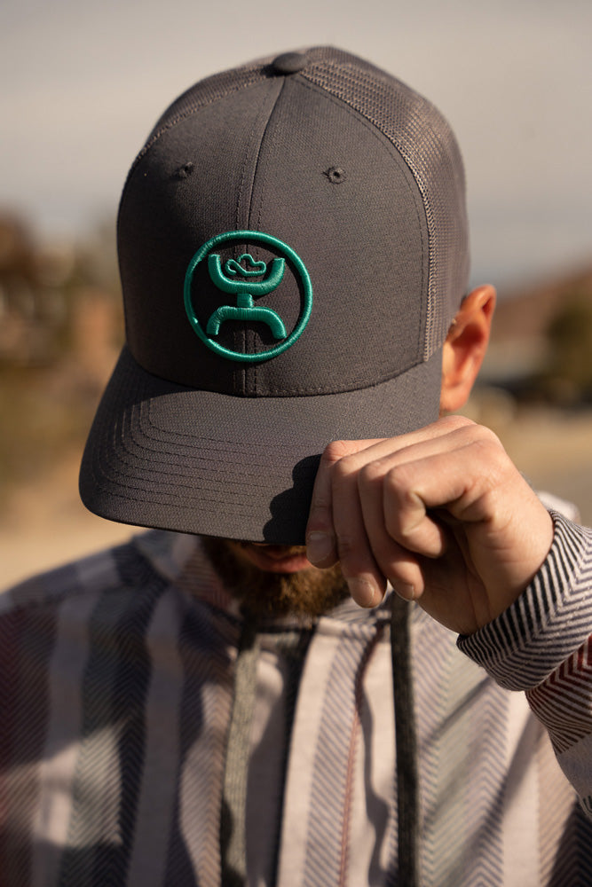 male model wearing mulit colored serape jacket with black and charcoal hat with teal logo