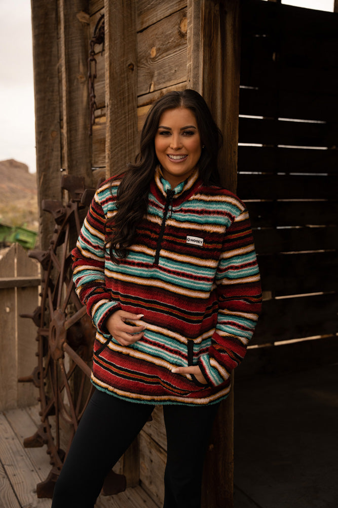 brunet, female model wearing dark jeans, red, blue, white serape pullover, posing in doorway of rustic wood building