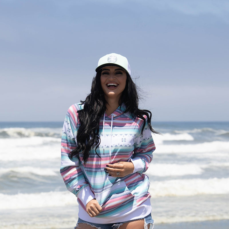 female model in denim shorts, white hooey hat and mesa hoody with green, teal, orange, red, yellow, white serape pattern in beach setting