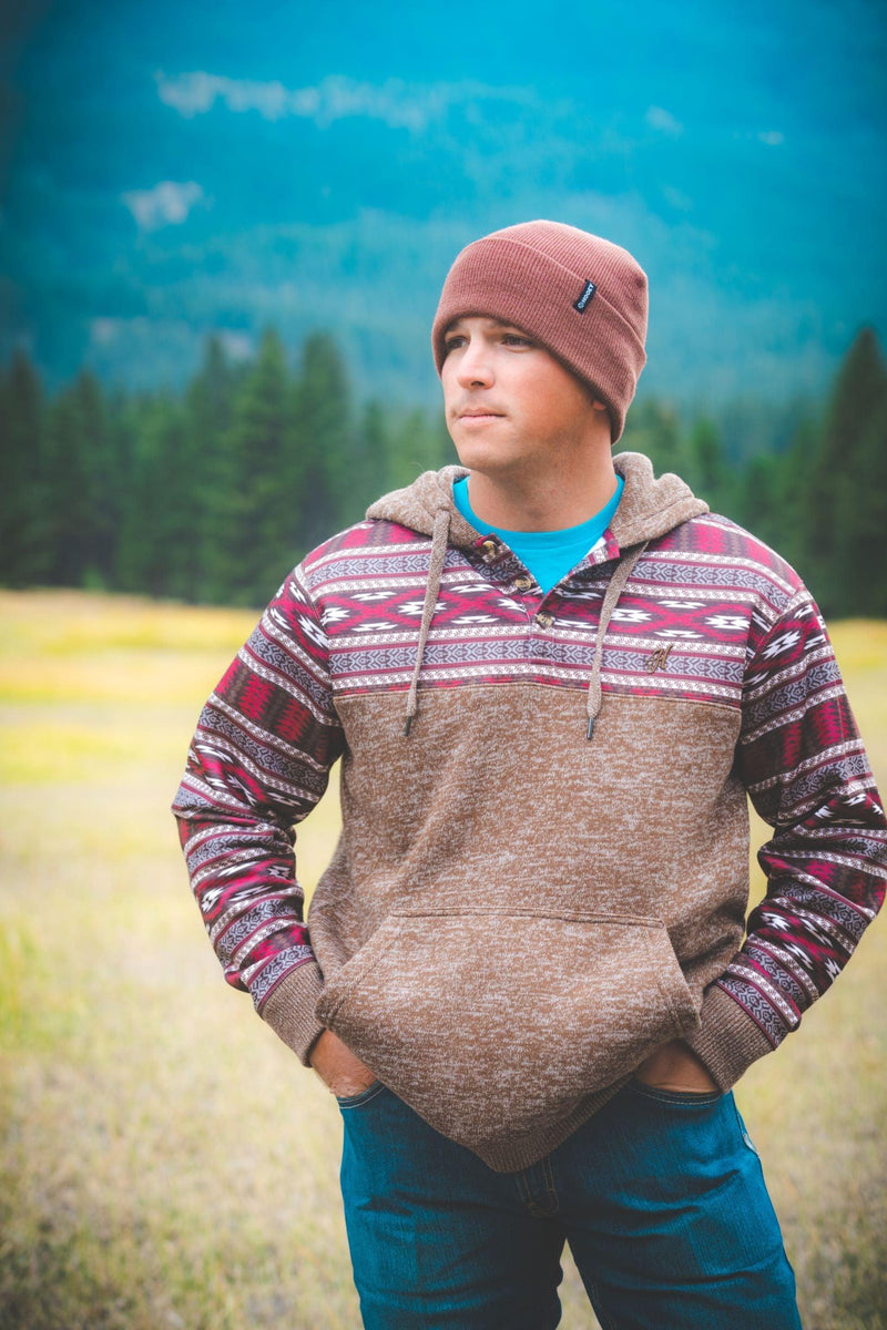 male model wearing the Morocco white hoody with tan, orange, blue, grey, purple patten on sleeves, pocket, and hood, jeans, and brown toboggan in outdoor setting