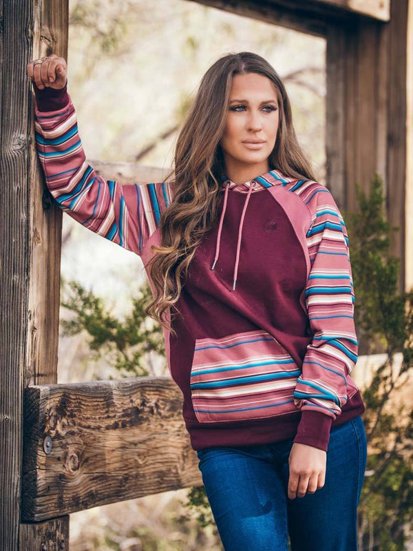 female model wearing the Savannah maroon hoody with red, tan, blue, maroon serape pattern on sleeves and hood and jeans near wood frame